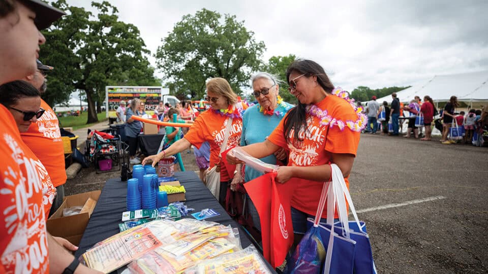 Three people in orange and light blue shirts look at swag items at a program table at Outreach at the Beach.