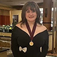 Young woman with dark hair and bangs wears a black dress and a medal around her neck.