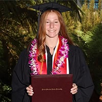 A young woman with red hair, a cap and gown and a lei around her neck holds a diploma.