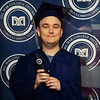 Young man dressed in navy blue cap and gown stands in front of navy blue backdrop with Del Mar College logos on it.