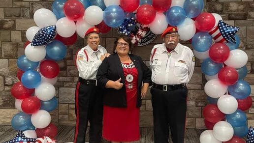 Council Member Jennifer Woods poses for a photo with two members of the Choctaw Honor Guard in front of a red white and blue balloon arch.