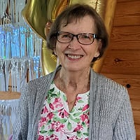 Older woman with short brown hair, wearing glasses, a grey cardigan and a shirt with pink and green flowers on it, smiles.