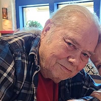 Older man with grey hair, wearing a red t-shirt with a blue flannel over it smiles.