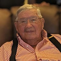 An older man in a gingham pattern shirt and suspenders smiles.