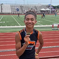 A young man standing on track holds up a medal as he smiles.