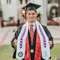 Young man, in cap and gown, proudly shows off his Choctaw Graduation Stole.