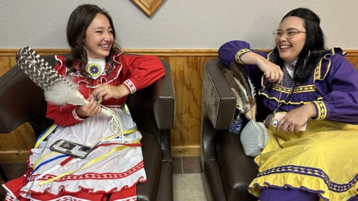 Tow young women in traditional choctaw dresses smile as they speak to each other backstage of the 2023 Princess Pageant.