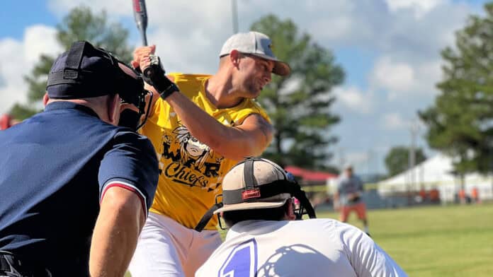 A baseball player in a yellow jersey and a gray ball cap swings at the baseball as an umpire and catcher waits for the pitch.