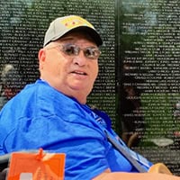 Older man in a blue shirt, a ball cap and sunglasses sits in front of a war memorial wall.