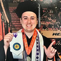 Young man in doctorate cap and gown hold Choctaw Graduation in one hand and makes OSU pistols firing sign with the other.