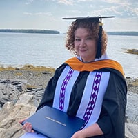 A woman with curly red hair, wearing a cap and gown, sits on the rocks beside a large body of water, holding a diploma.