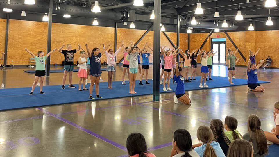 A group of cheerleaders practice their moves while a group of girls watch them at a CNO Summer Camp.