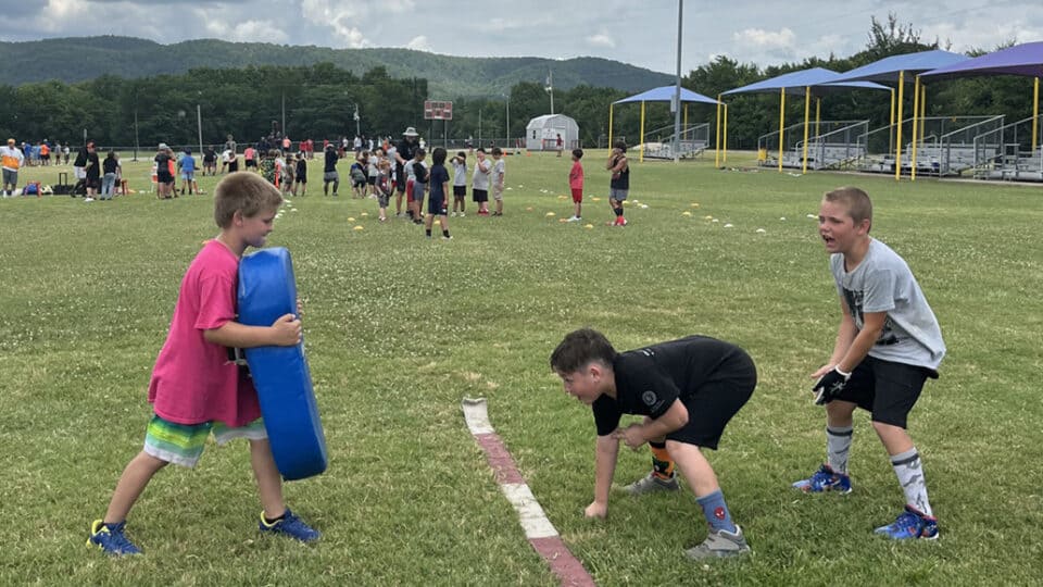 A boy in a red shirt holds a pad for a boy in a black shirt to practice his football tackle at a CNO Summer Camp.