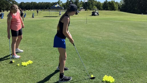 A girl in a red shirt watches a girl in a black shirt practice her golf put at a CNO Summer Camp.