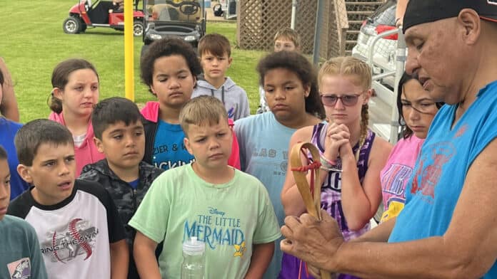 A man holds a stickball stick while a group of kids look at it with wonder in their eyes at a CNO Summer Camp.