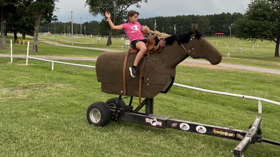 A young girl rides a mechanical horse with her hand in the air at a CNO Summer Camp.