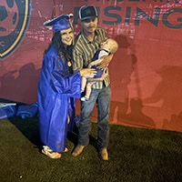 Brook Boehne in a blue cap and gown smiles for a photo with a young man holding a baby.