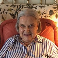 Eugenia Miller, an older woman sits on an orange chair and smiles for the camera.