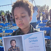 Logan Porter, young boy with brown curly hair looks down as he holds a photo of himself.