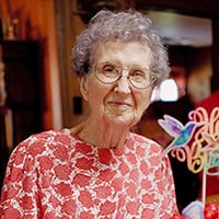 Stella Heemer, older woman with short gray hair and glasses, dressed in red and white floral shirt smiles.