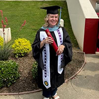 Stephane Peterson, a woman with blonde hair, dressed in a cap and gown, smiles as she holds a red rolled-up piece of paper.