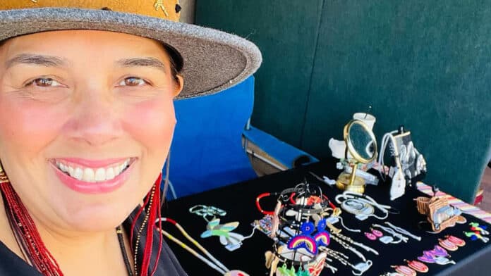 Deana Ward, a woman in a wide-brimmed hat and red earrings poses for a photo in front of a booth with beadwork on it.