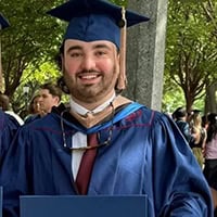 Weston Duffy, young man with a short beard dressed in a blue cap and gown smiles at the camera.