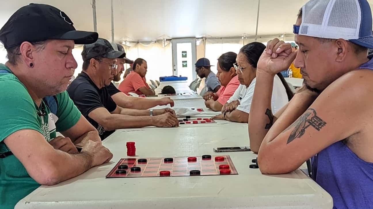 A man in a green shirt and a man in a purple shirt look down at their checkerboard during a game of checkers.