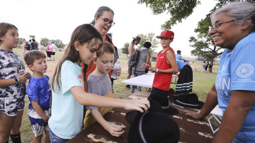 Jessa Taylor lifts the hat to correctly guess where the prize was hidden as her brother, Jacob, inspects it during the hat game demonstration in the Choctaw Village during the 2024 Labor Day festival