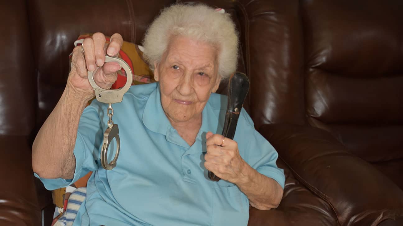 An old woman in a blue blouse holds up handcuffs and a flashlight.