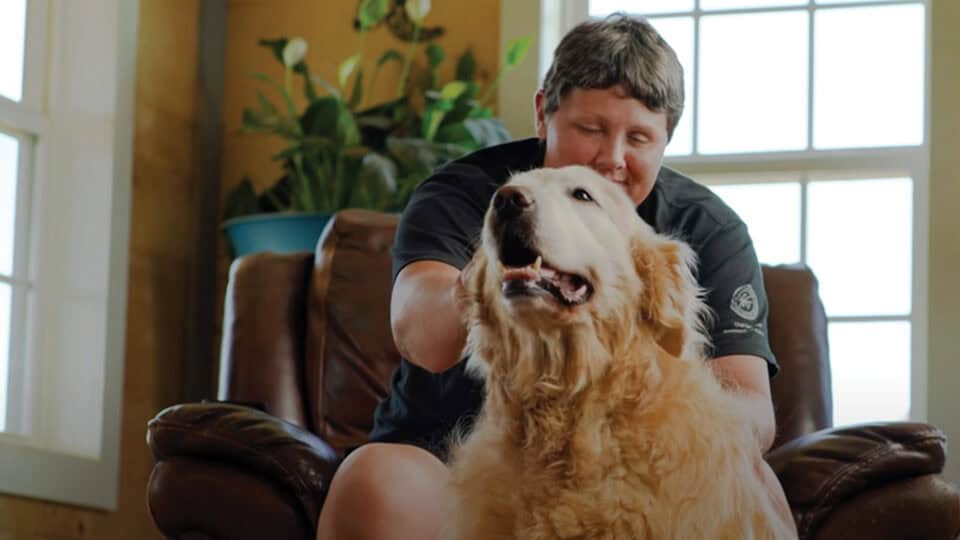 Woman with short hair sits on a leather sofa chair while petting a golden retreaiver.