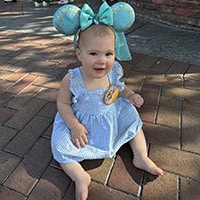 Toddler in blue dress and turquoise Mickey Mouse ears sets on the bricked ground.