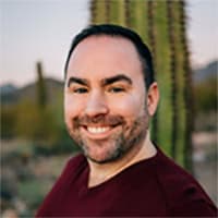 A man wearing a maroon t-shirt smiles for a photo in front of saguaro cacti.