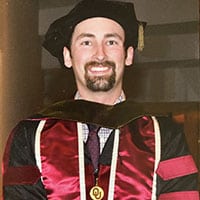 A young man with a goatee, wearing a doctoral gown, smiles for the camera.