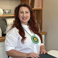 A woman with red hair and a white button-up shirt sits at a desk, wearing a Choctaw seal medallion.