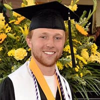 A young man in a cap and gown smiles for a photo in front of a bouquet of yellow flowers.