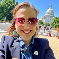 A young woman in sunglasses and a navy blue sports coat smiles for a selfie in front of the Capitol building.