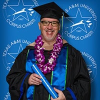 A man in a cap and gown, wearing black-rimmed glasses and a purple lei, stands against a blue background with Texas A&M University-Corpus Christi logos.