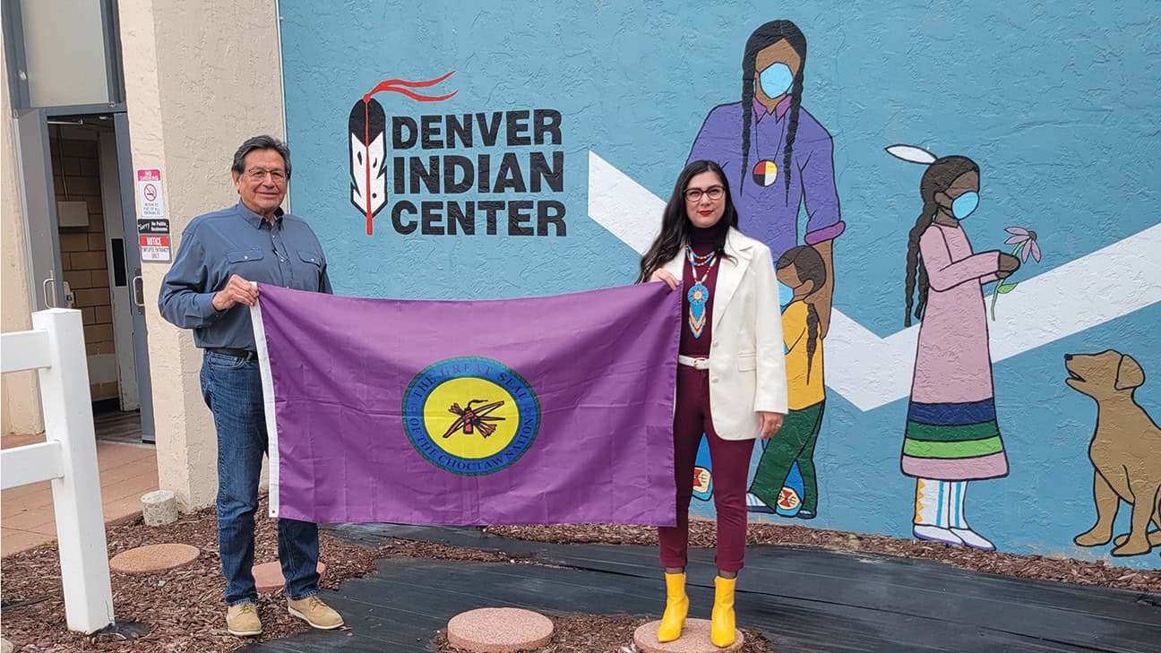 Two people stand in front of the Denver Indian Center holding the Choctaw Nation of Oklahoma flag