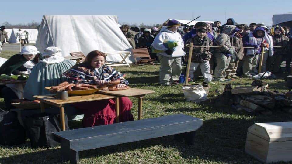 Raven Baker, in 1800’s style Choctaw dress, cuts sausage in preparation for the day’s meal. Behind are visitors of the park interacting with students involved in a cooking demonstration.