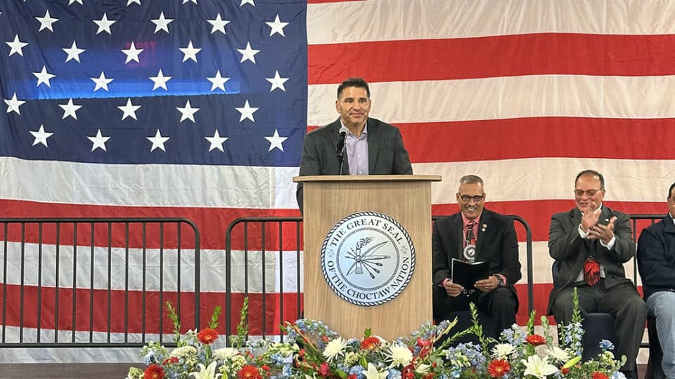 Man speaks at podium in front of a large American flag, and the Chief and Assistant Chief smile in the background.