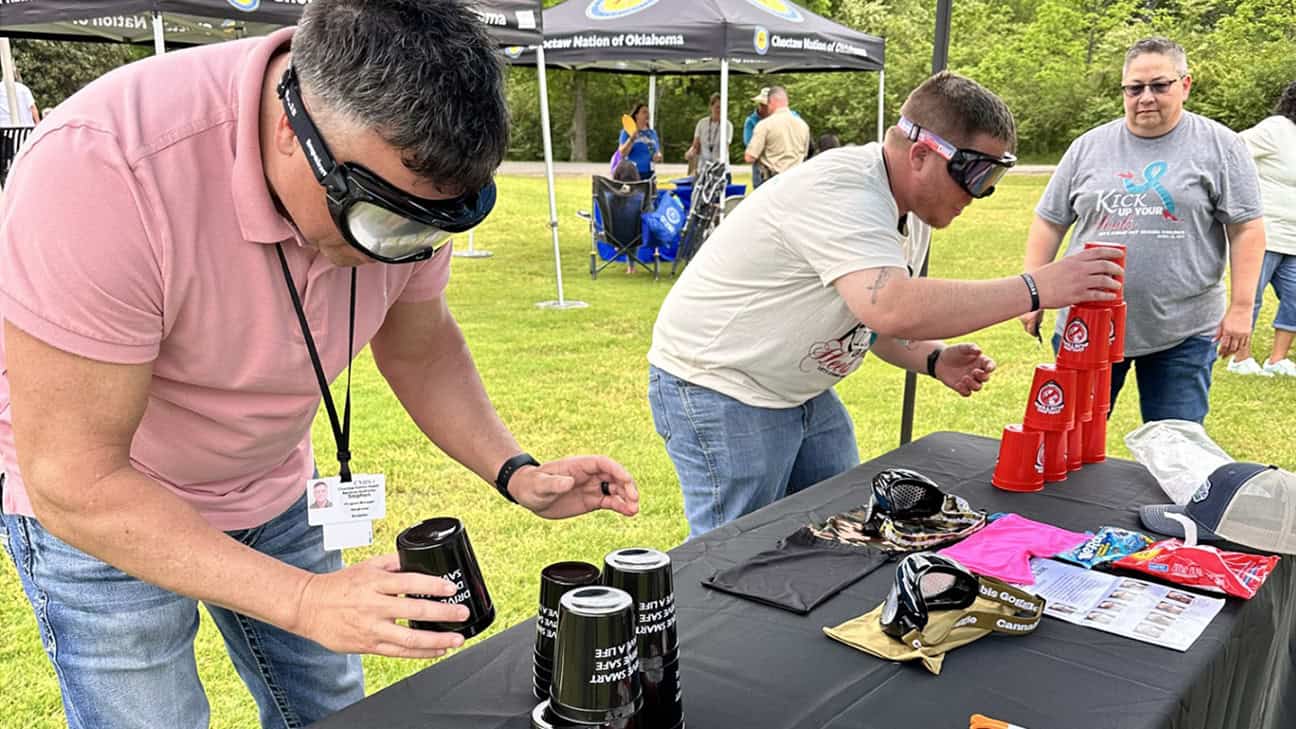 Two men attempt to stack cups while wearing Drunk Buster goggles.