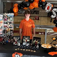 A young man in an orange shirt stands in front of a table filled with photos and desserts.