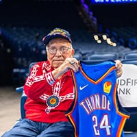 Elder dressed in Choctaw traditional shirt hold up OKC Thunder jersey.