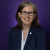 A woman with glasses, wearing a business suit, smiles in front of the purple backdrop.