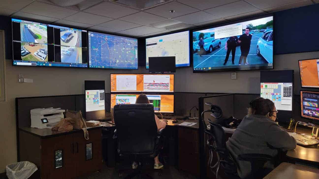 Two women sit in a room full of tv and computer screens, answering phones.