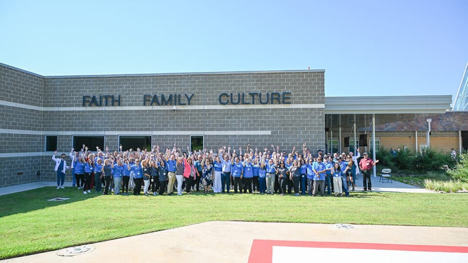 A large crowd waves to the camera in front of a building with faith, family and culture on it.