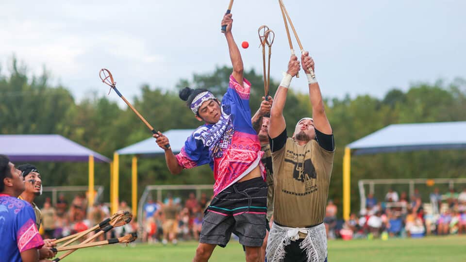 Two men jump towards teh ball in an aggressive stickball game.