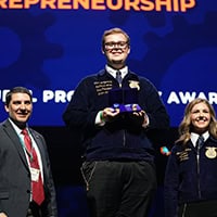 A young man with glasses, dressed in an FFA uniform, holds up the award on stage.