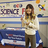 A young girl holds up a trophy in front of a sign that says science fair.